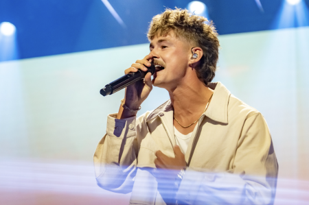 "Male singer with mullet performs on stage, holding a mic, wearing a beige jacket, with vibrant blue and white stage lighting in the background."