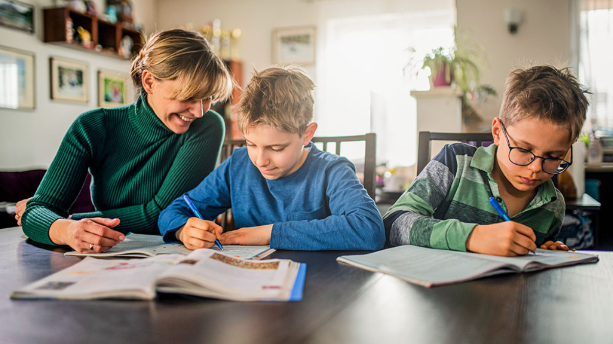 A woman and two boys using a Homeschool Daily Learning Tracker at a table, writing in books, in a bright and cozy home setting.