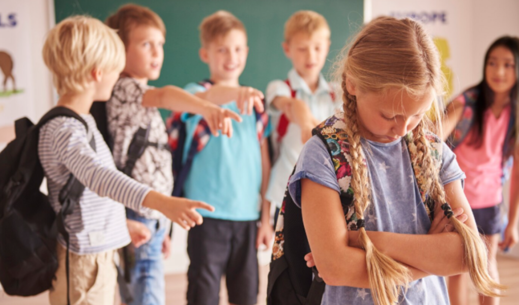 A young girl looks sad while a group of boys point and laugh at her in a classroom, illustrating one of the social problems in the education system.