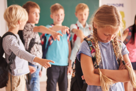 A young girl looks sad while a group of boys point and laugh at her in a classroom, illustrating one of the social problems in the education system.