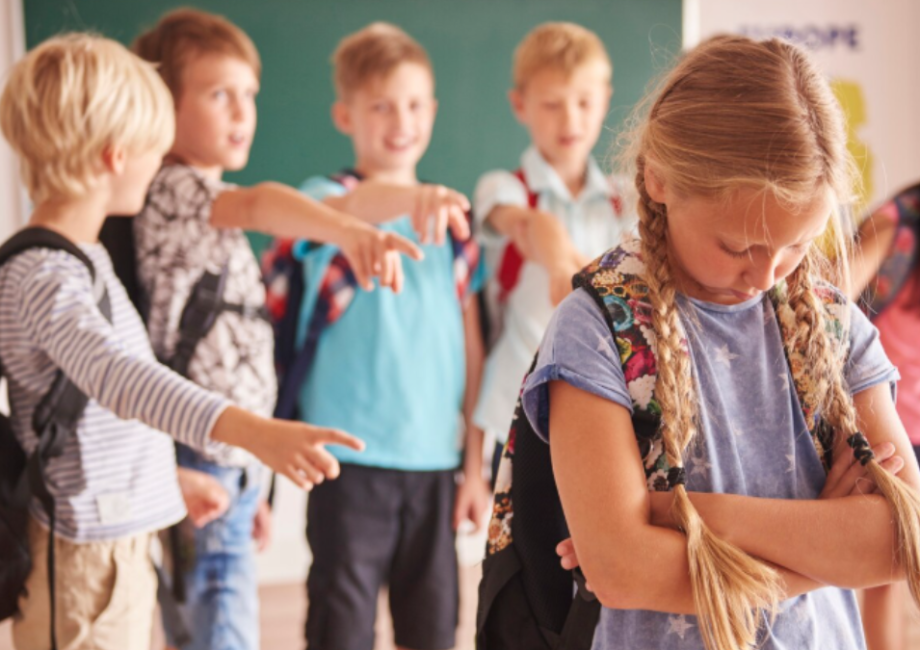 A young girl looks sad while a group of boys point and laugh at her in a classroom, illustrating one of the social problems in the education system.